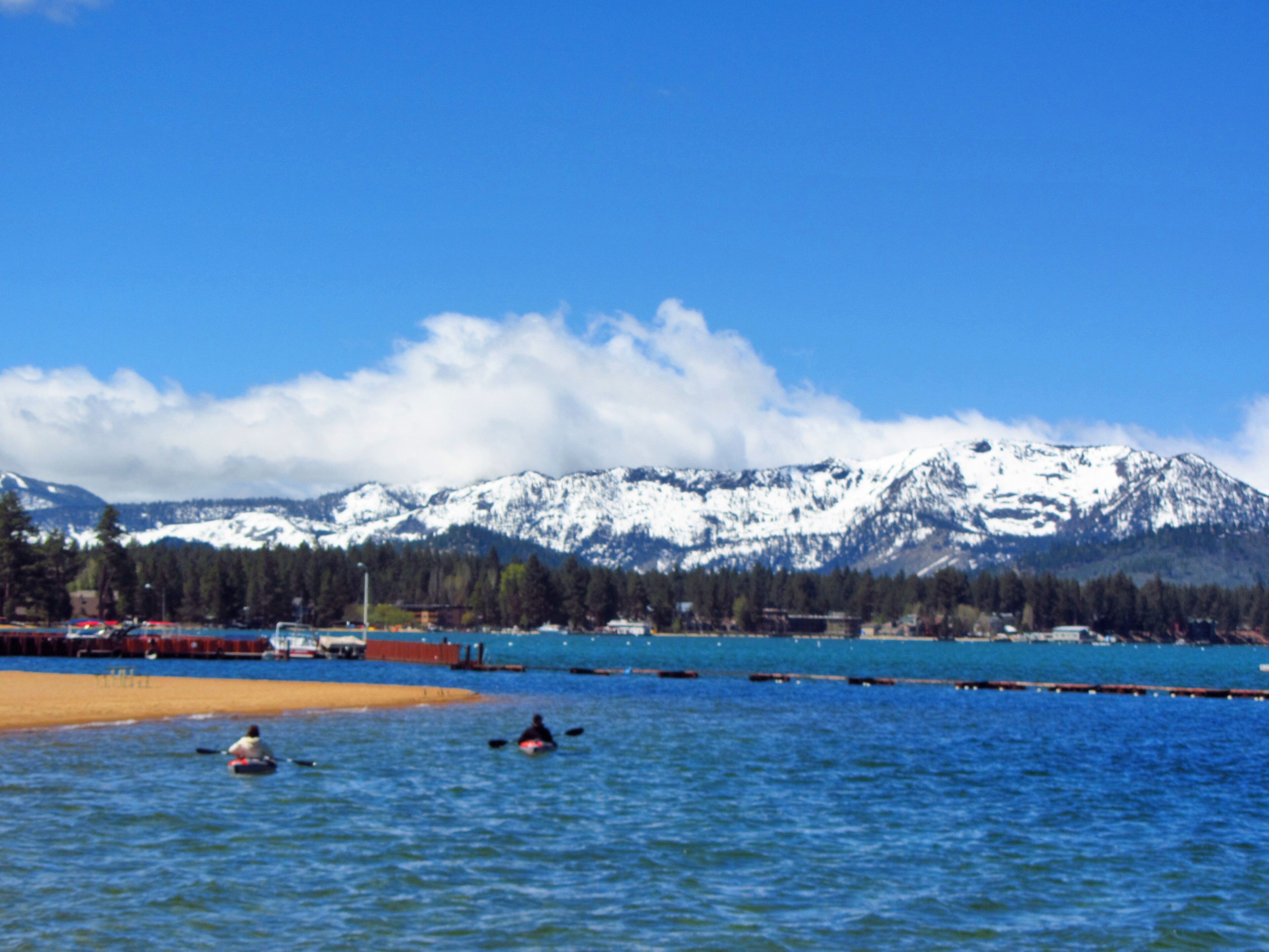 Un lago con kayakistas, una playa arenosa y montañas nevadas al fondo.