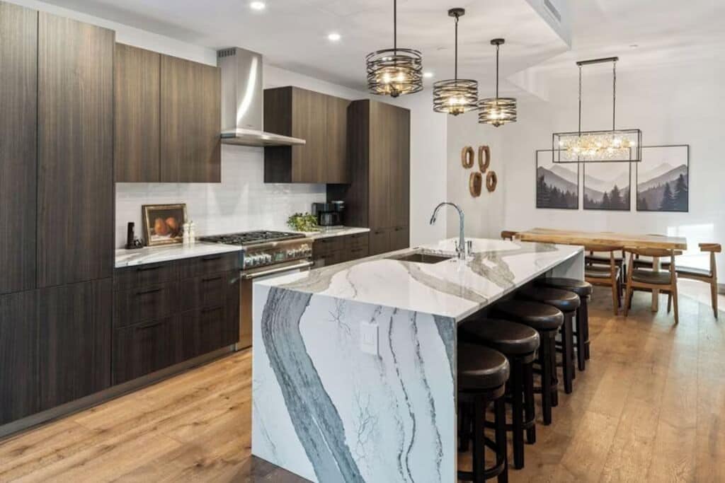 A modern kitchen with dark wood cabinets, a marble island, bar stools, pendant lighting, and a dining area with a wooden table and chairs.