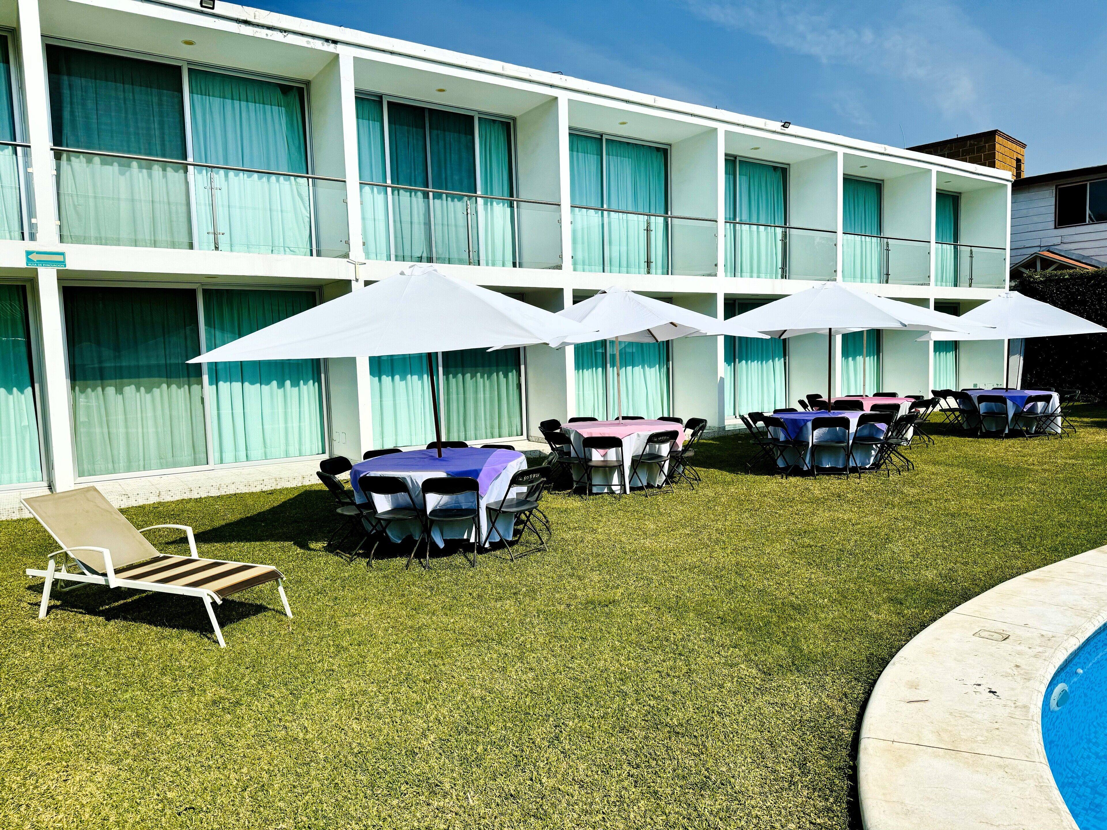A hotel courtyard with outdoor seating and umbrellas, a swimming pool, and a building with large windows and curtains.