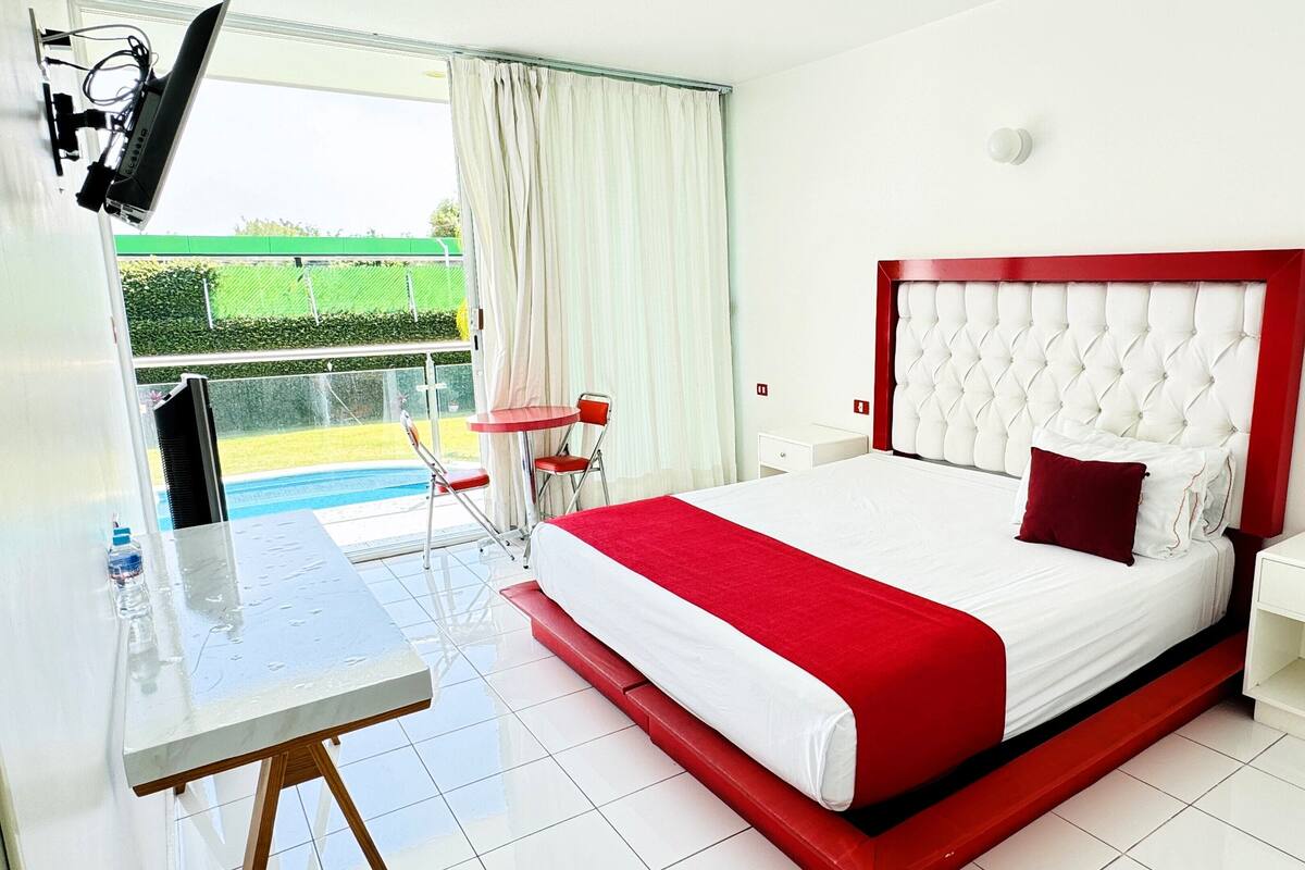 A hotel room with a large bed, a small table, a chair, and a view of a swimming pool.