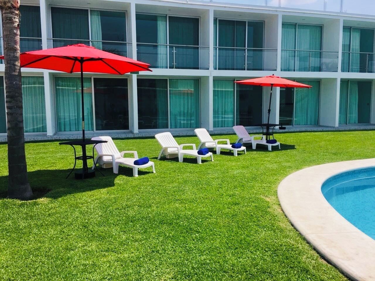 A poolside area with white lounge chairs and red umbrellas, a palm tree, and a building with large windows in the background.