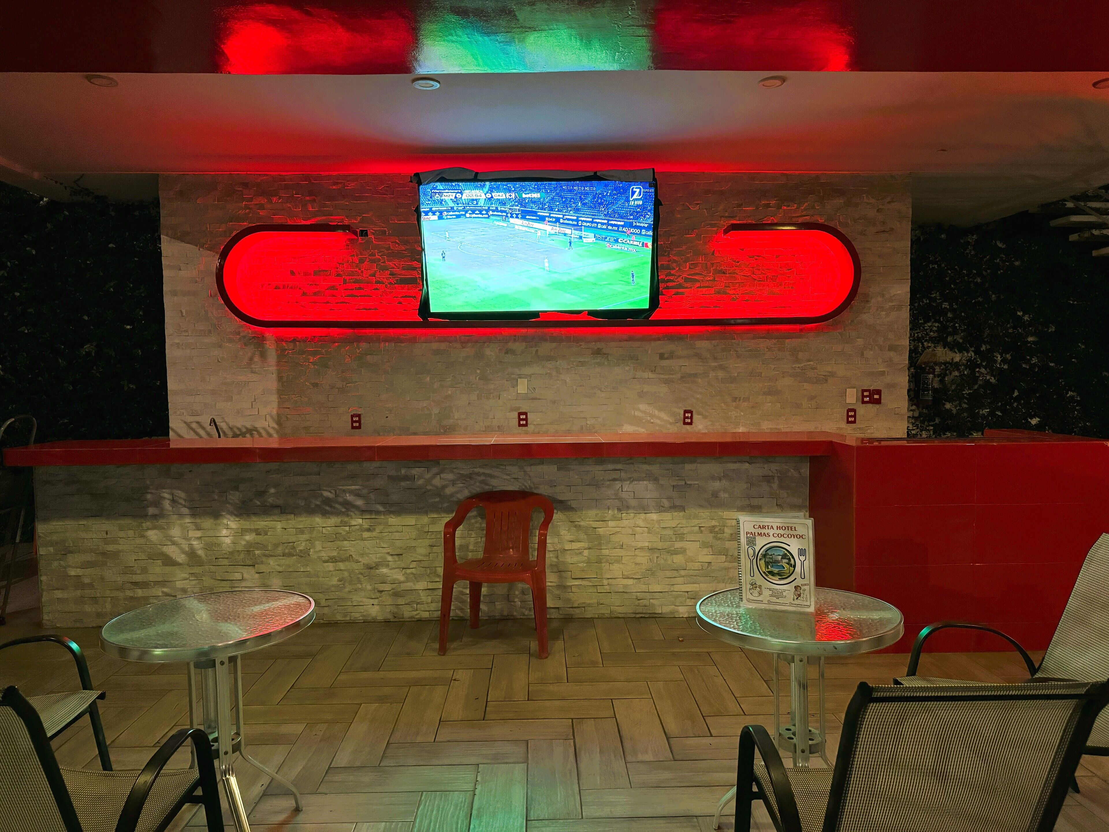 A hotel lobby with a television mounted on a brick wall, a wooden chair, and two small tables with chairs.