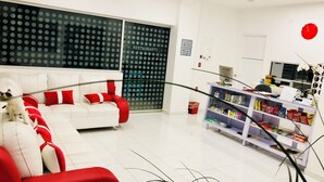 Un salon moderne avec un canapé blanc et des coussins rouges, une cloison en verre ornée d’un motif à pois, et une étagère à provisions contenant divers produits emballés.