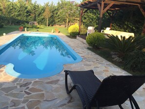 A swimming pool with stone tiles, a wicker chair, and a pergola in a garden setting. - Villa Oneiro (Preveza)
