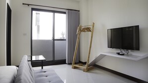 Une chambre d’hôtel moderne équipée d’un canapé, d’un téléviseur fixé au mur, d’un porte-manteau en bois et d’une porte-fenêtre coulissante donnant sur un espace extérieur.