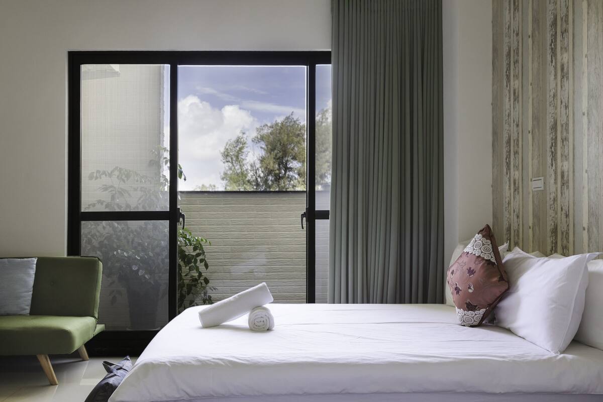 Ein Hotelzimmer mit einem Bett, einem grünen Sessel und Blick auf Bäume durch ein großes Fenster.
