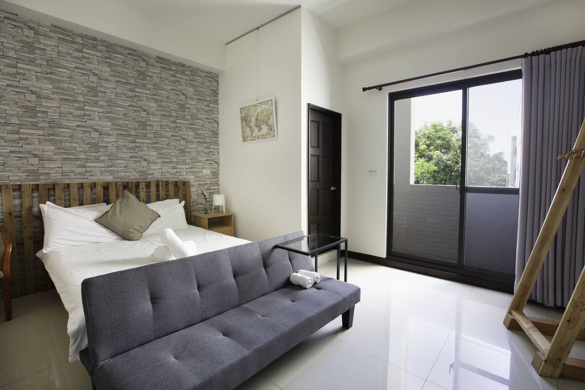 Ein modernes Schlafzimmer mit grauem Sofa, Holz-Kopfende, Bett mit weißer Bettwäsche, einer Weltkarte an der Wand und einem großen Fenster mit Vorhängen.