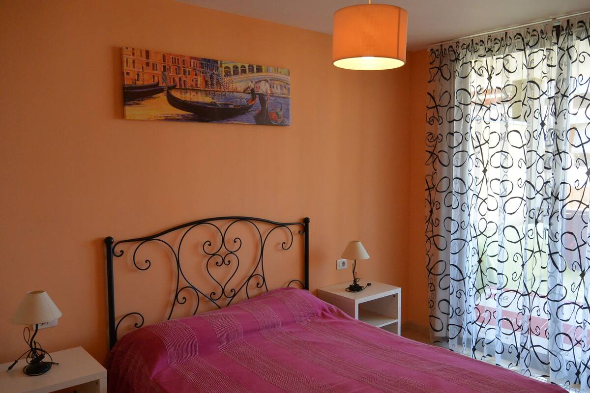 A bedroom with a bed, a nightstand with a lamp, a painting of a canal scene, and patterned curtains.