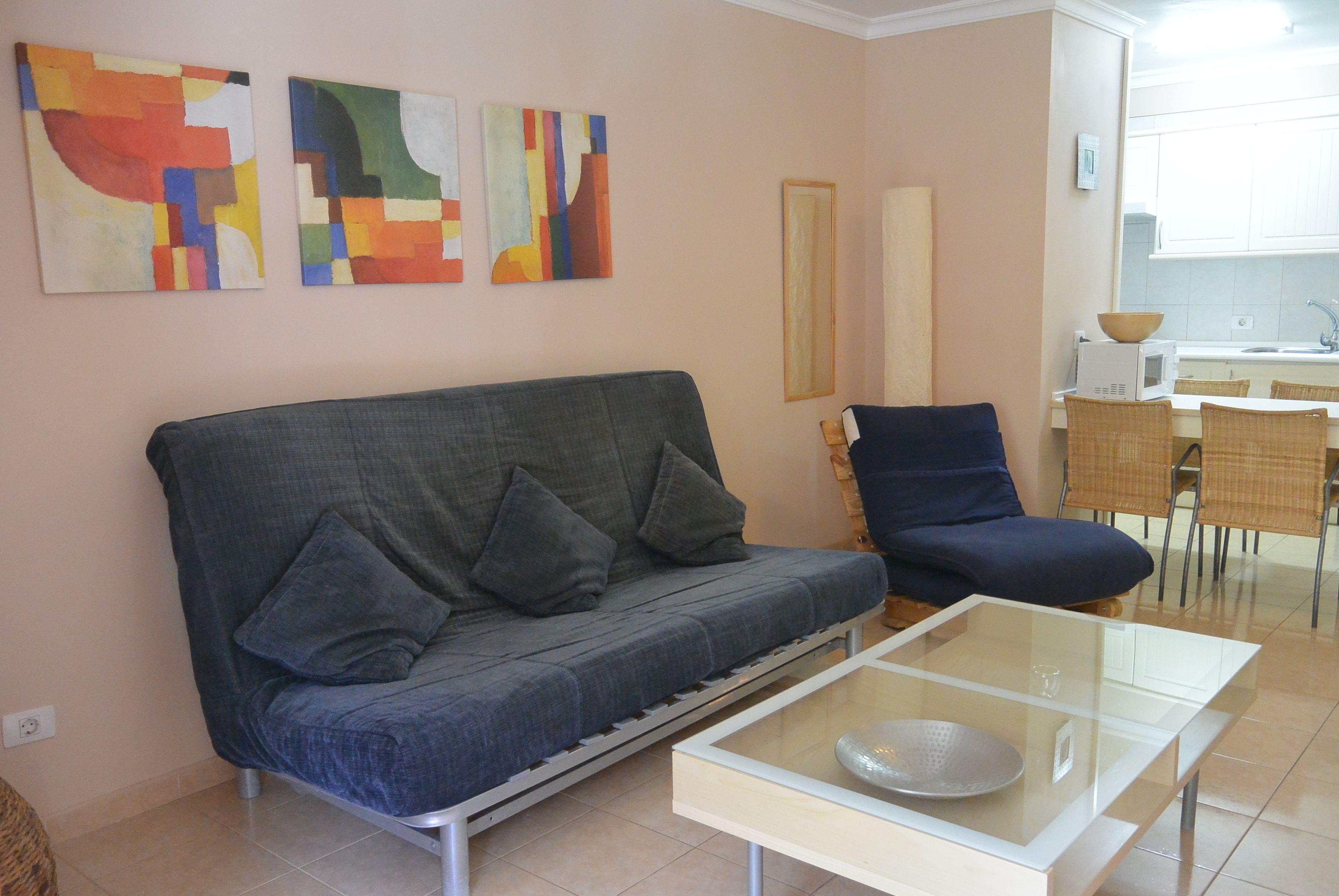 Ein Wohnzimmer mit grauem Sofa, einem Glastisch und einem blauen Sessel. An den Wänden befinden sich abstrakte Gemälde, im Hintergrund ist eine Küchenzeile erkennbar.