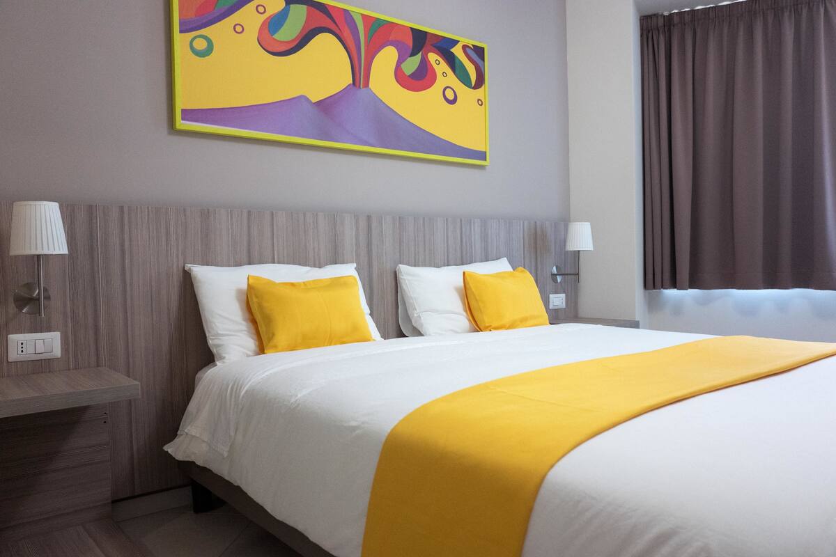 A hotel room with a large bed, two bedside lamps, a wall-mounted artwork, and a window with curtains.