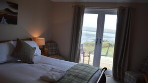 Ein Hotelzimmer mit Bett, Sessel, Lampe und Meerblick durch eine Glastür.