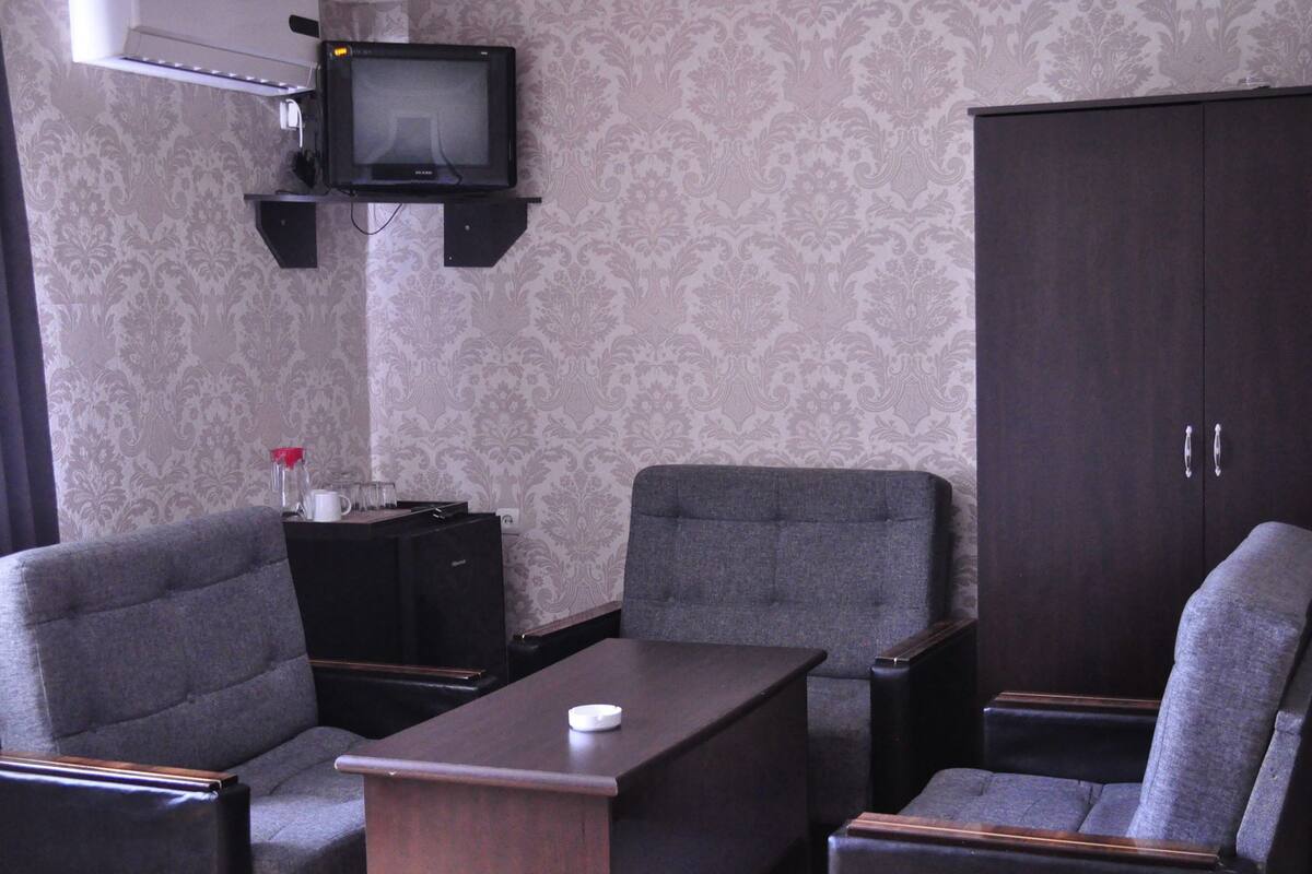 A room with two grey armchairs, a small table with a white object, a dark wooden cabinet, and a mounted television.