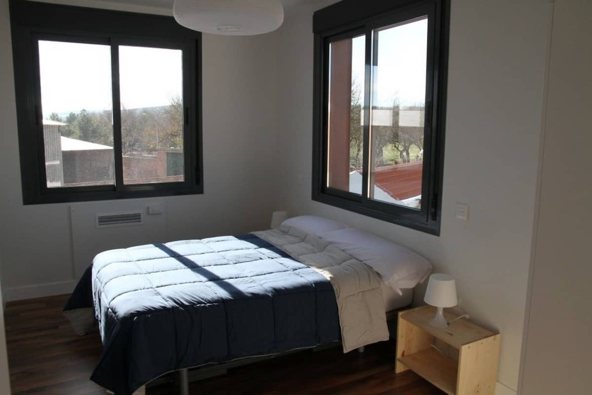 Ein Schlafzimmer mit einem Bett, zwei Fenstern, einem Nachttisch und einer Lampe.