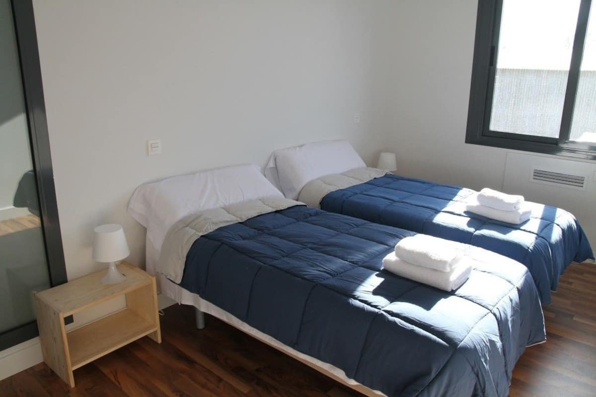 Ein Hotelzimmer mit zwei Betten, jedes mit einer blauen Bettdecke und weißen Kissen. Ein kleiner Nachttisch mit einer Lampe steht daneben, und ein Fenster lässt Tageslicht herein.