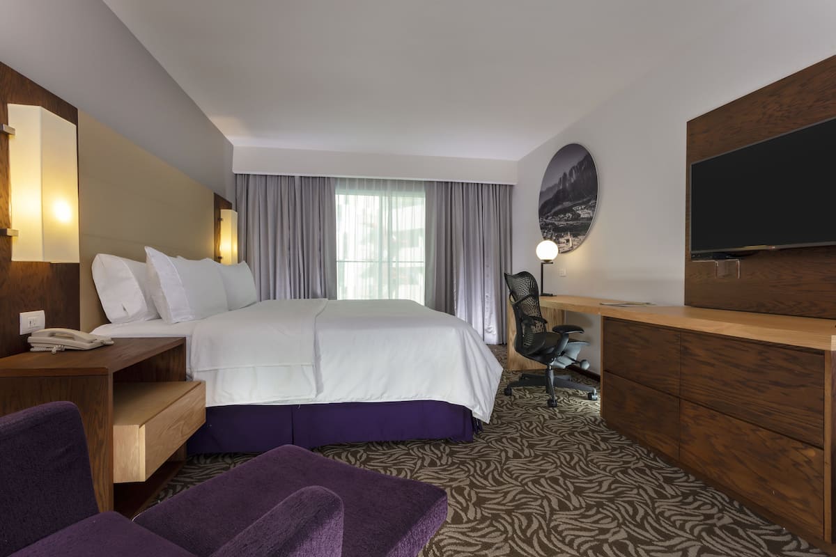 Habitación de hotel con una cama grande, un escritorio con silla, televisor y un ventanal con cortinas.