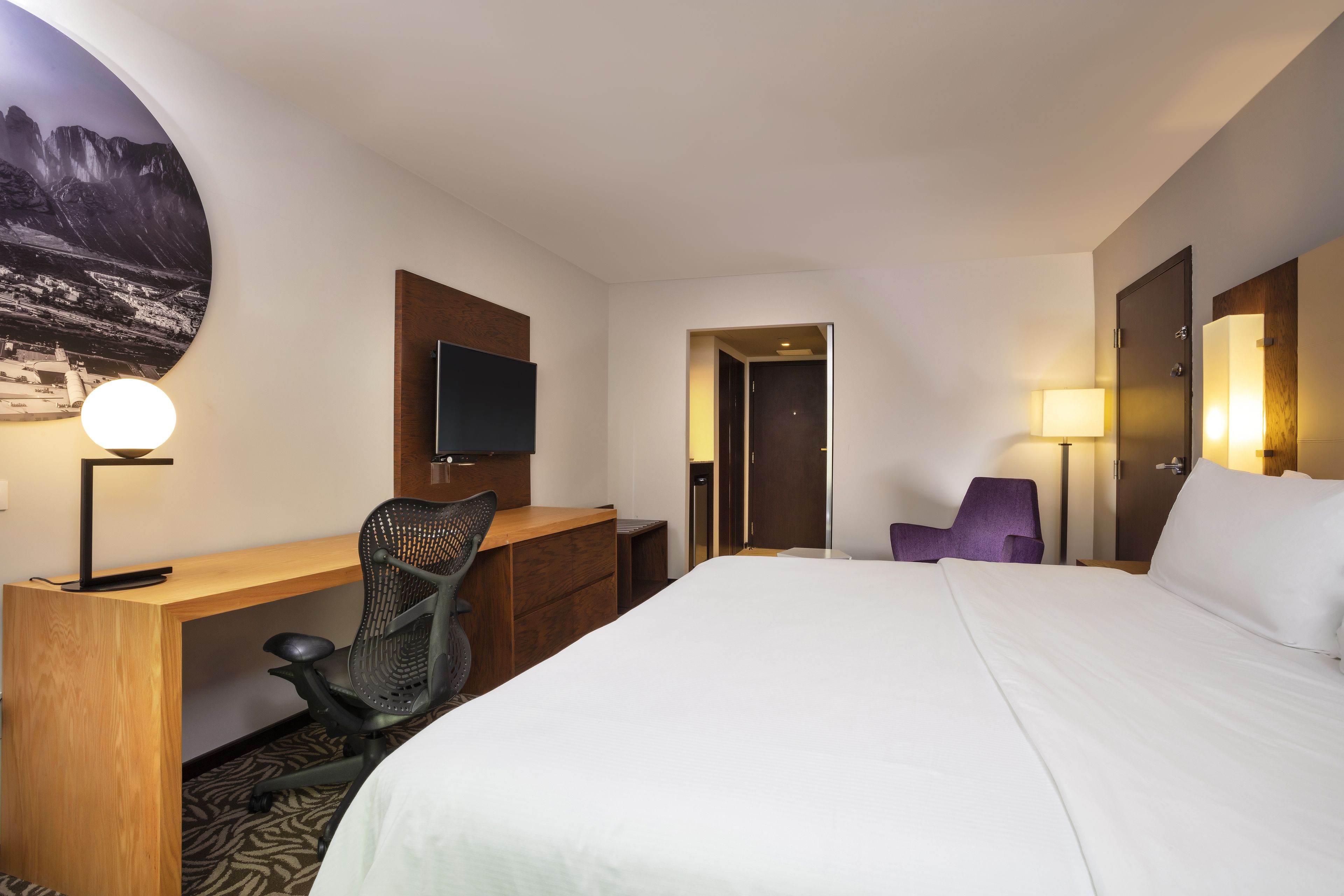 Habitación de hotel con cama, escritorio, silla, televisor y una puerta que da a otra habitación.