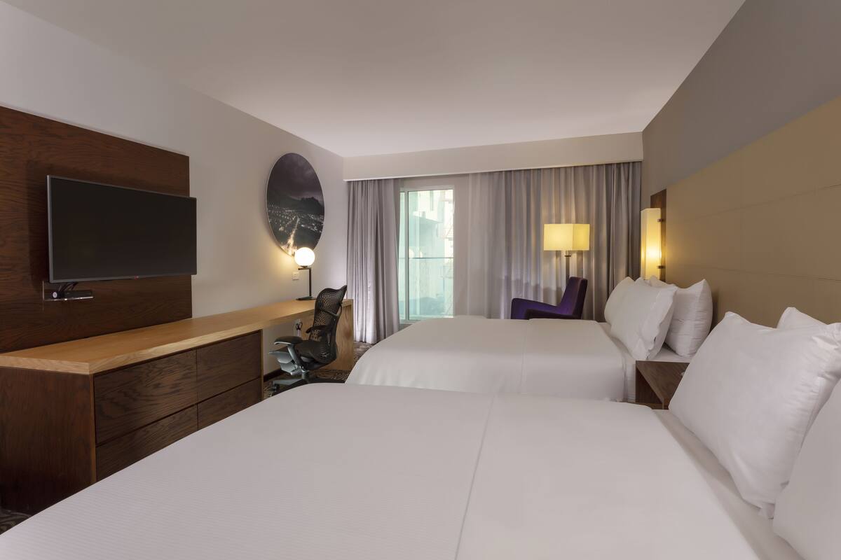 Habitación de hotel con dos camas, una televisión de pantalla plana, un escritorio con una silla, una lámpara y vistas a la ciudad a través de amplias ventanas.