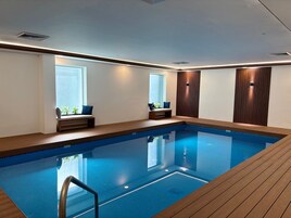 Una piscina interior moderna con muelle de madera, asientos integrados y grandes ventanales.
