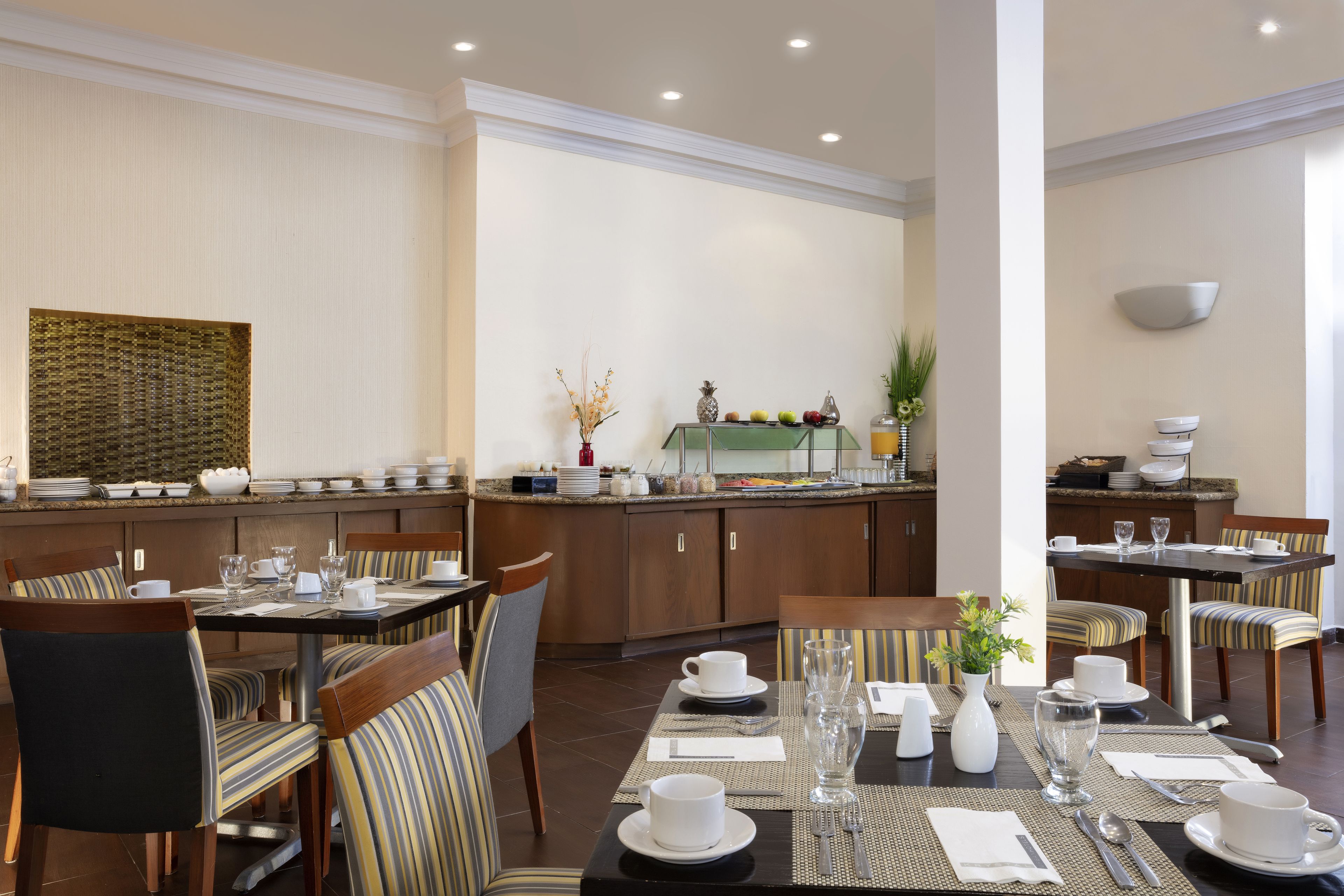 Une salle à manger bien éclairée, avec plusieurs tables préparées pour le petit-déjeuner, où l’on propose une variété de plats et de boissons.