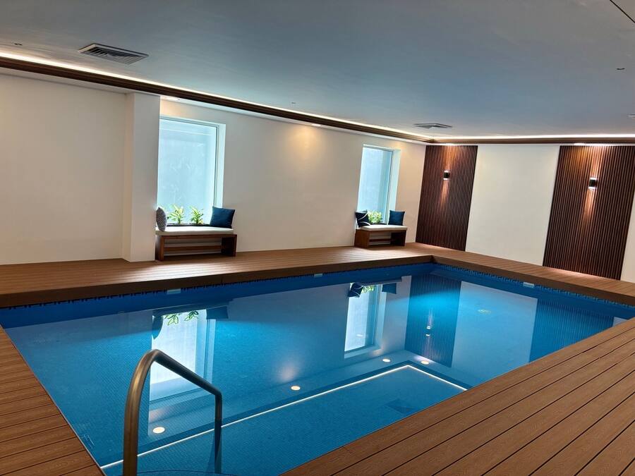 Una piscina interior moderna con terraza y zonas de estar de madera, ventanas amplias y un diseño limpio y minimalista.