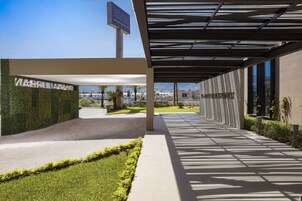 Una entrada de hotel moderna con un paseo techado, vegetación y una vista despejada del entorno.