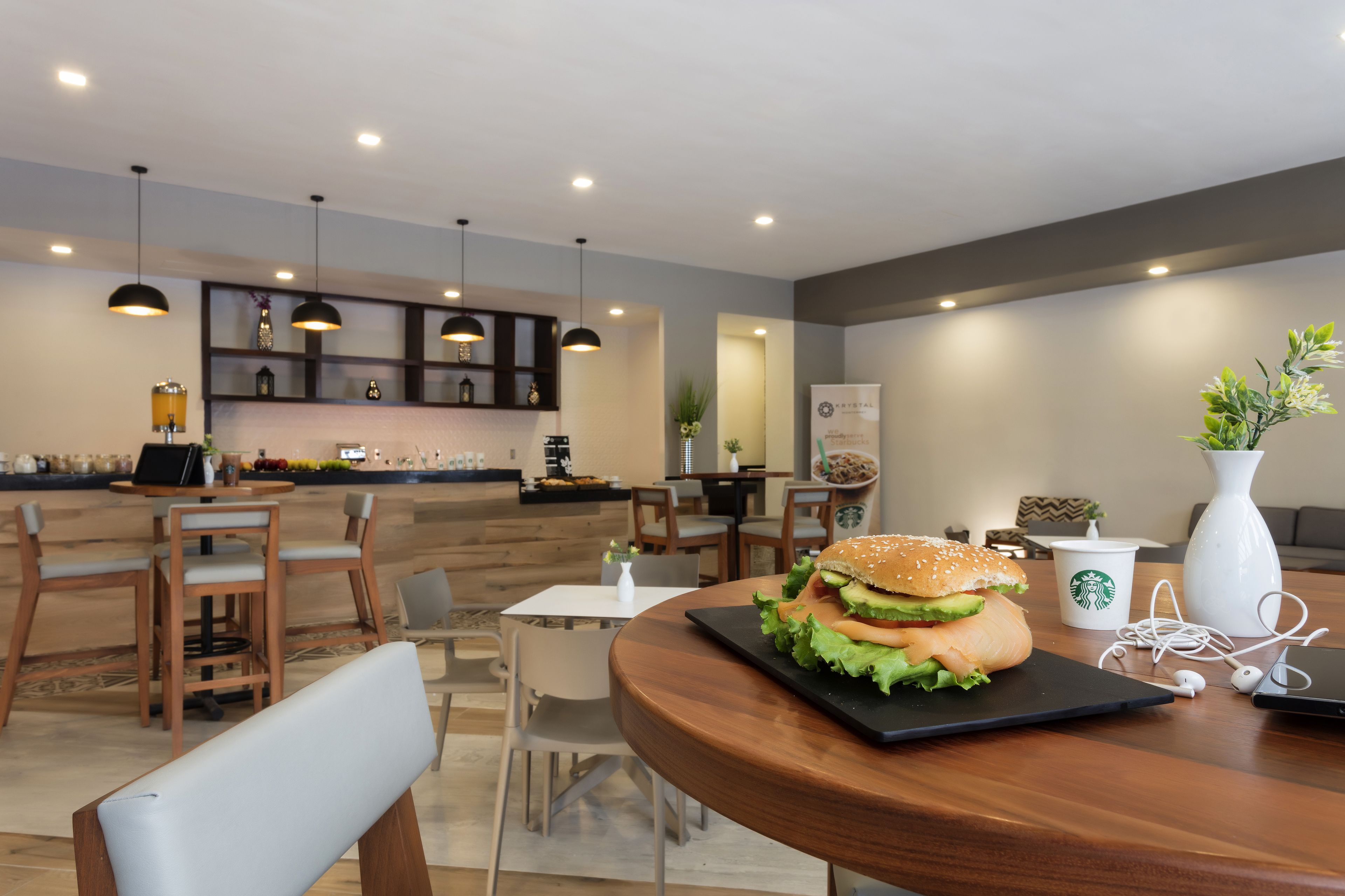 A modern dining area with wooden tables and chairs, a large burger on a black plate, a Starbucks cup, and a white vase with plants.