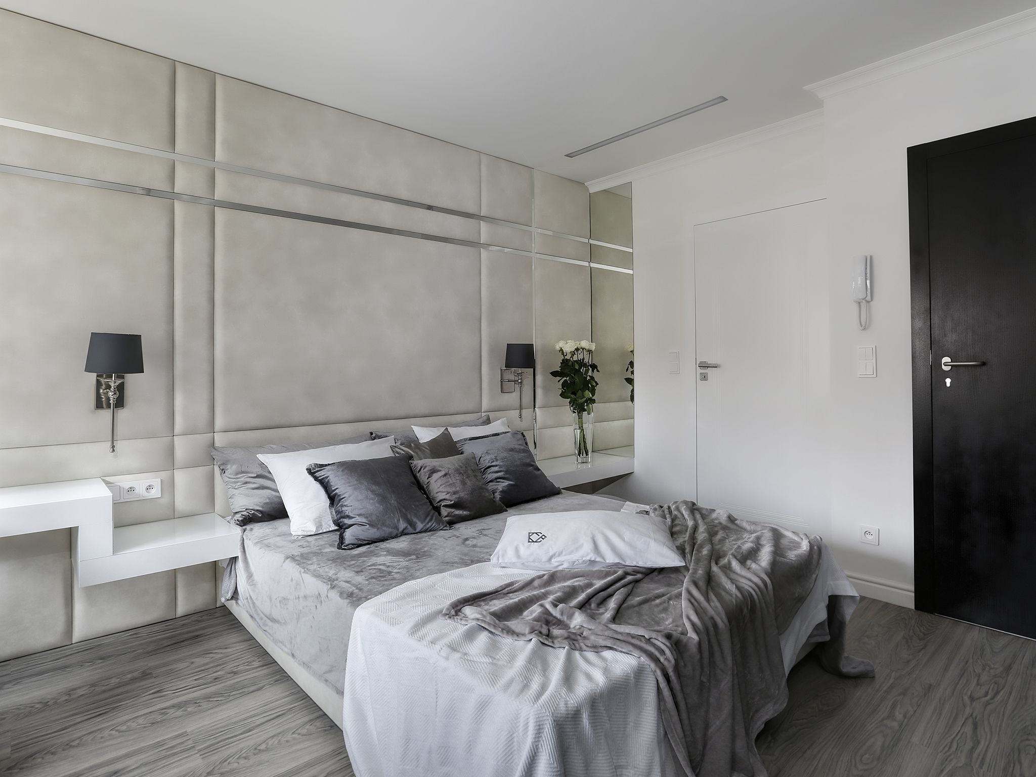 A modern bedroom with a large bed, bedside tables, a dark door, and a mirror on the wall.