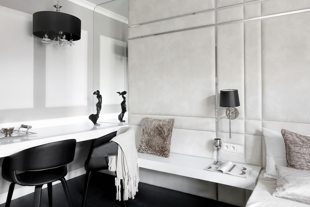 A modern hotel room with a white vanity, black chairs, a sofa with a white throw, and a wall-mounted lamp.