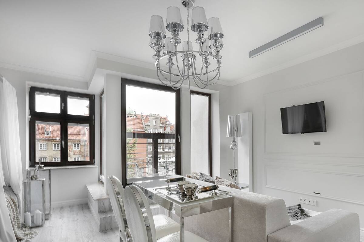 A modern dining area with a chandelier, a TV mounted on the wall, and a view of buildings outside.