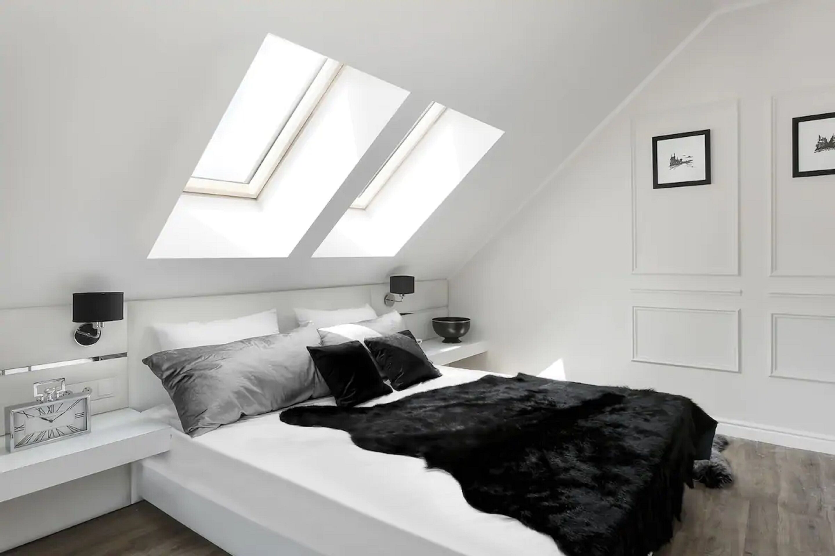 Une chambre moderne avec un puits de lumière, un lit avec des oreillers, une table de chevet avec une lampe et un mur orné de tableaux encadrés.