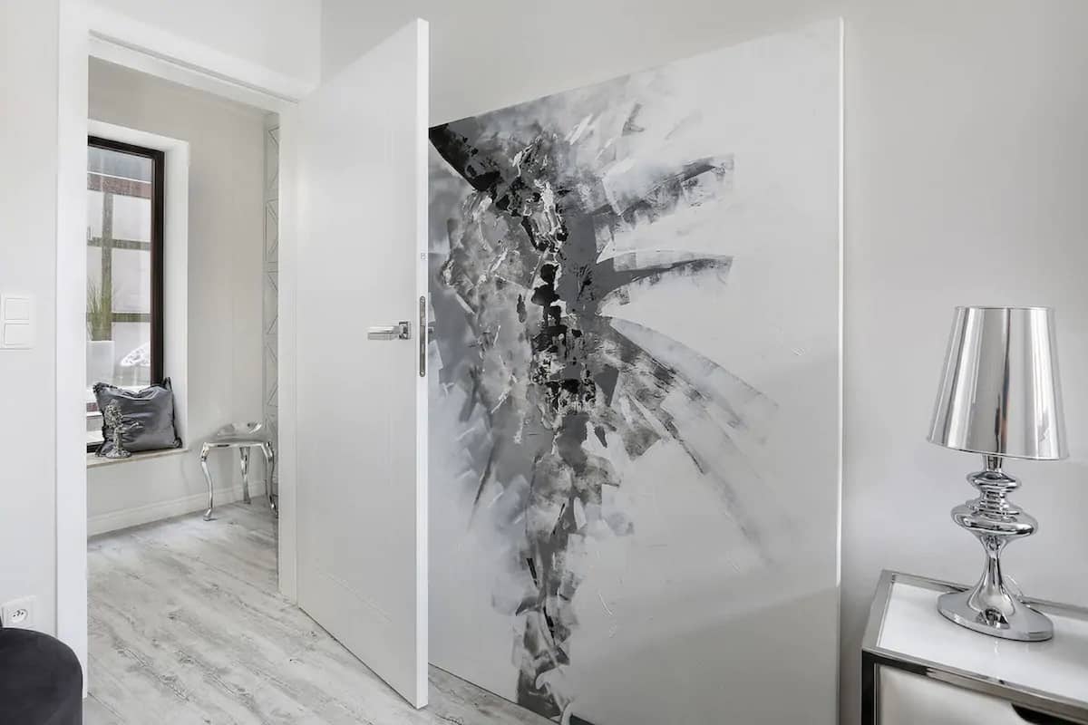 A modern room with a white door featuring a black and white abstract painting, a small table with a lamp, and a window with a view of the outside.