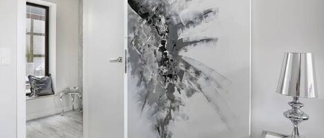 A modern room with a white door featuring a black and white abstract painting, a small table with a lamp, and a window with a view of the outside.