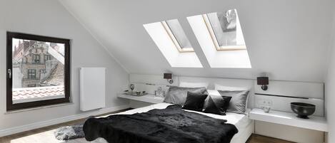 A modern bedroom with a skylight, a bed with black and white bedding, a bedside table with a lamp, and a view of buildings outside.