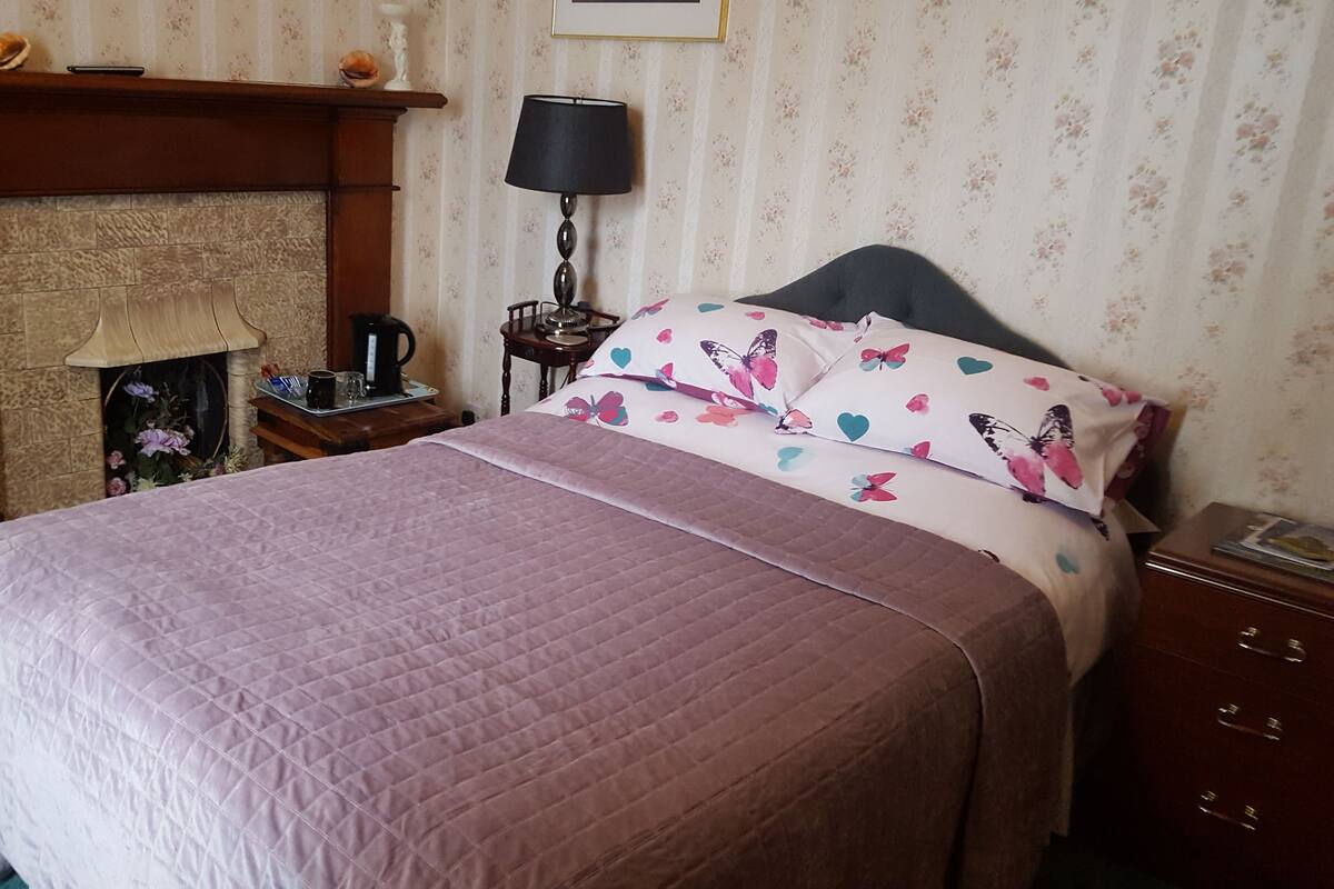 A neatly made bed with a floral pillow and a quilted blanket. A wooden nightstand with a lamp and a small table with a kettle and cups is beside the bed.