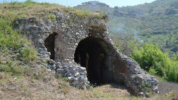 Estrutura de pedra com entrada em arco, rodeada por relva seca e arbustos, situada em paisagem montanhosa com um edifício ao longe.