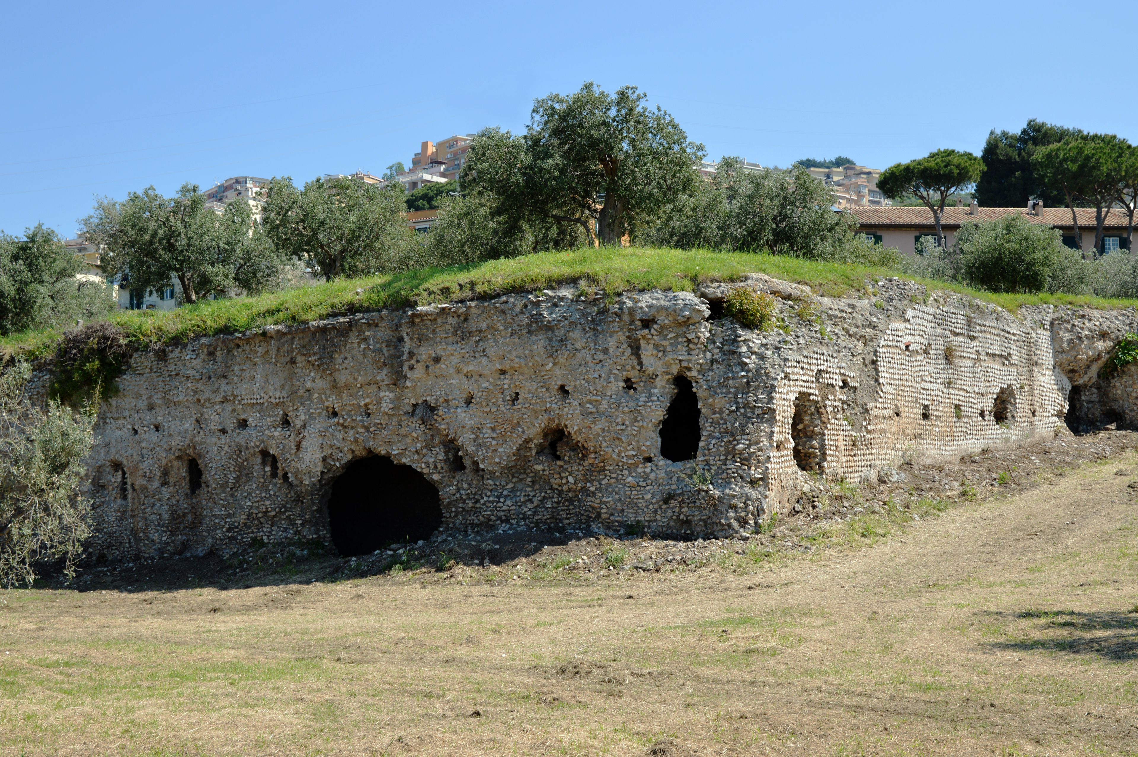 Una antigua estructura de piedra con entrada tipo túnel, rodeada de vegetación y edificios al fondo.