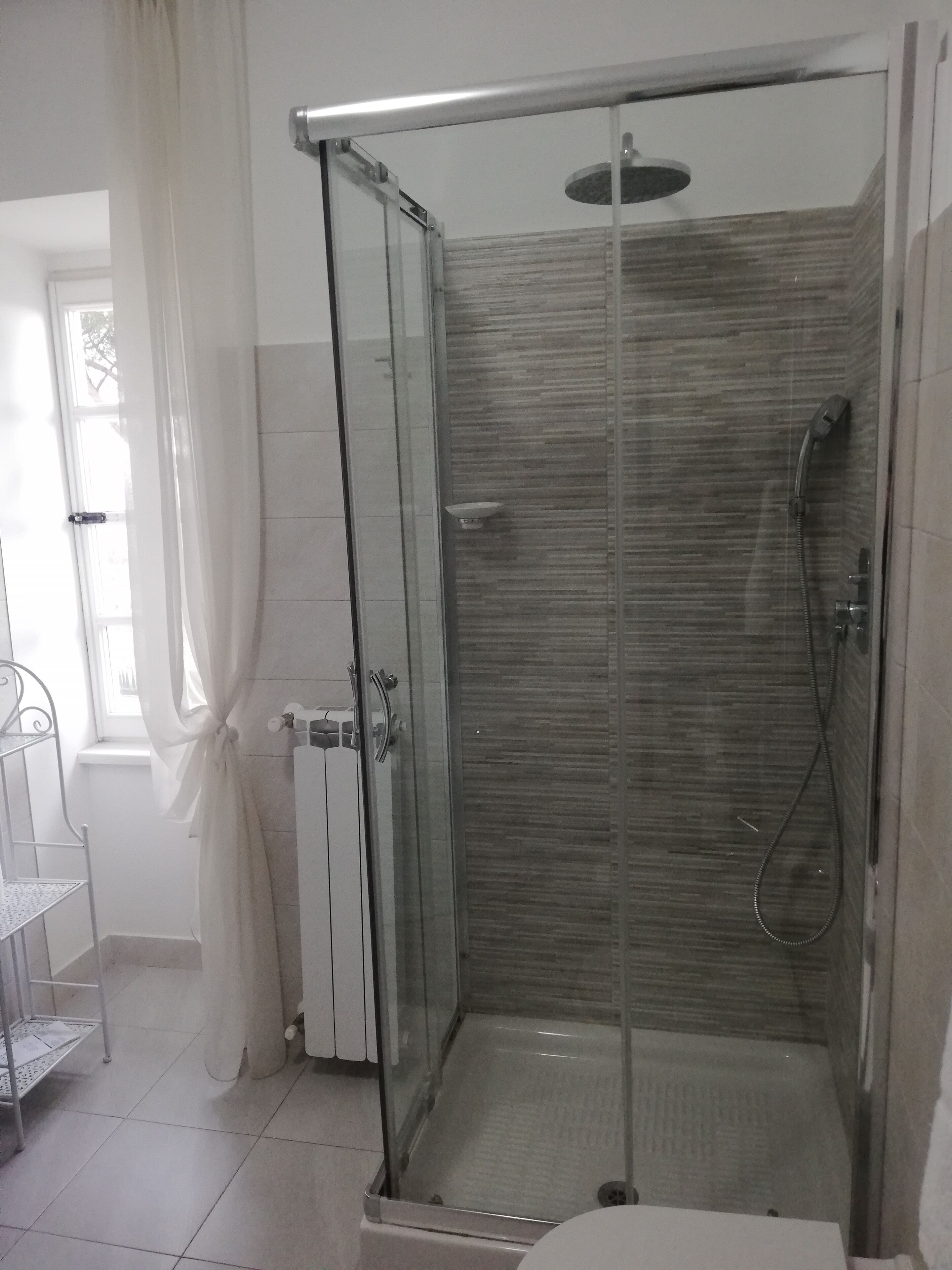 Een douche met glazen wanden, een betegelde vloer, een witte radiator en een metalen plankunit.