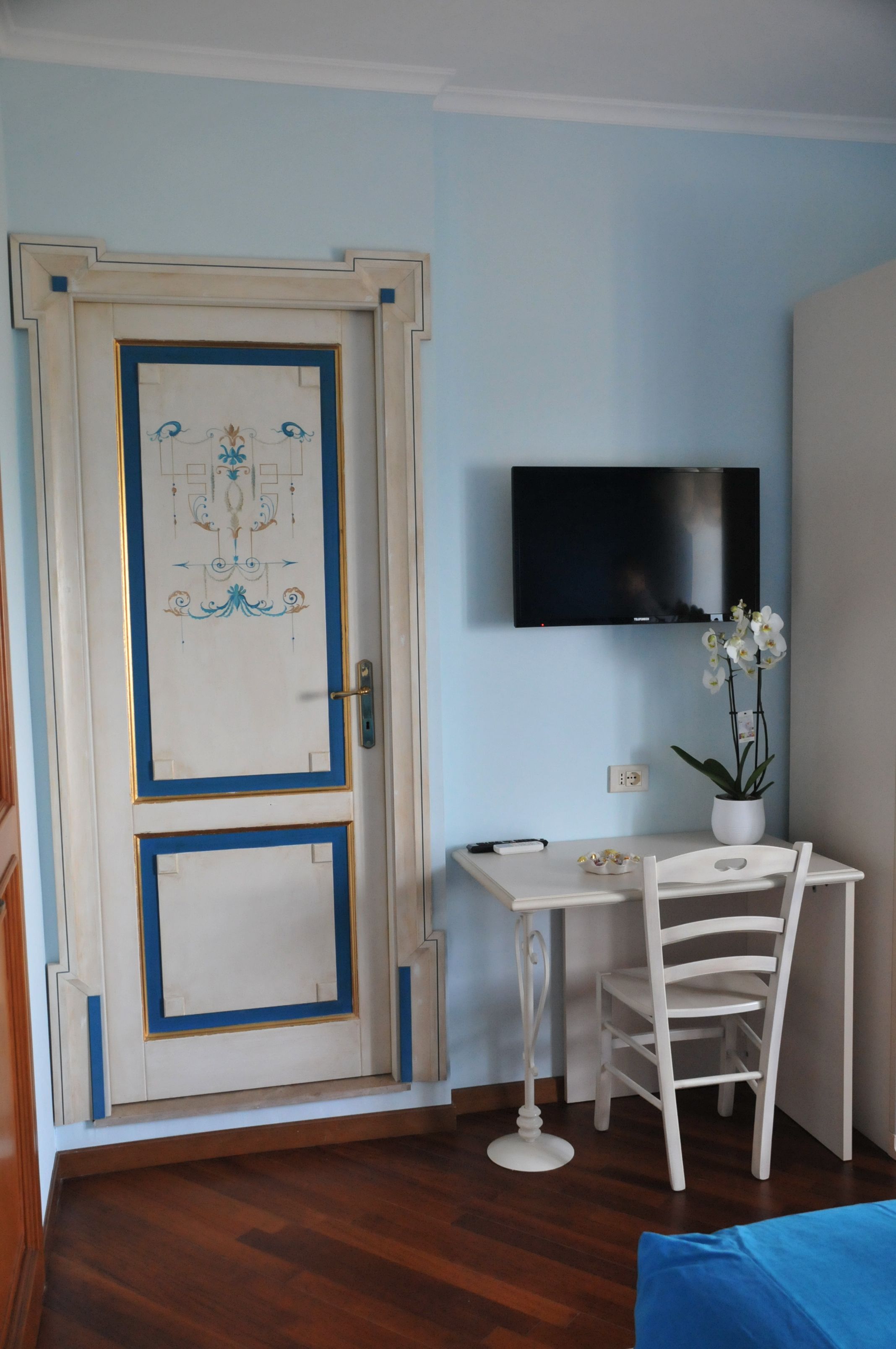 Una habitación con piso de madera, una puerta blanca con detalles azules, una pequeña mesa blanca con una silla, una orquídea en maceta y un televisor montado.