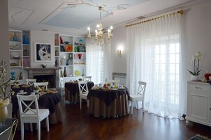 Un comedor con mesas redondas preparadas para el desayuno, sillas blancas, una lámpara de araña y una chimenea.