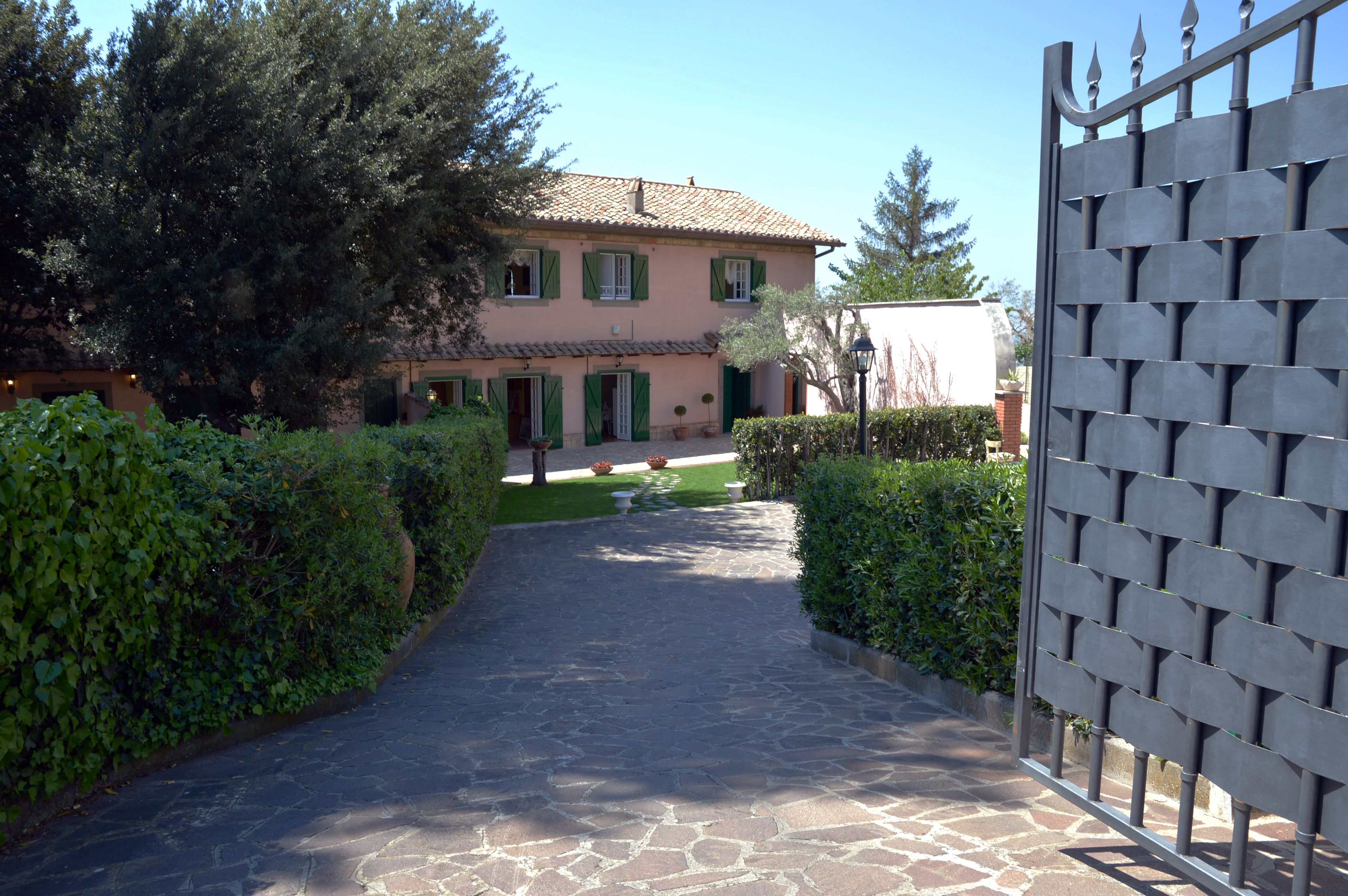 A stone pathway leading to a pink building with green shutters, surrounded by hedges and trees.