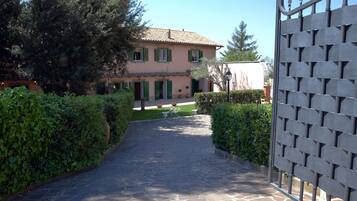 A stone pathway leading to a pink building with green shutters, surrounded by hedges and trees.