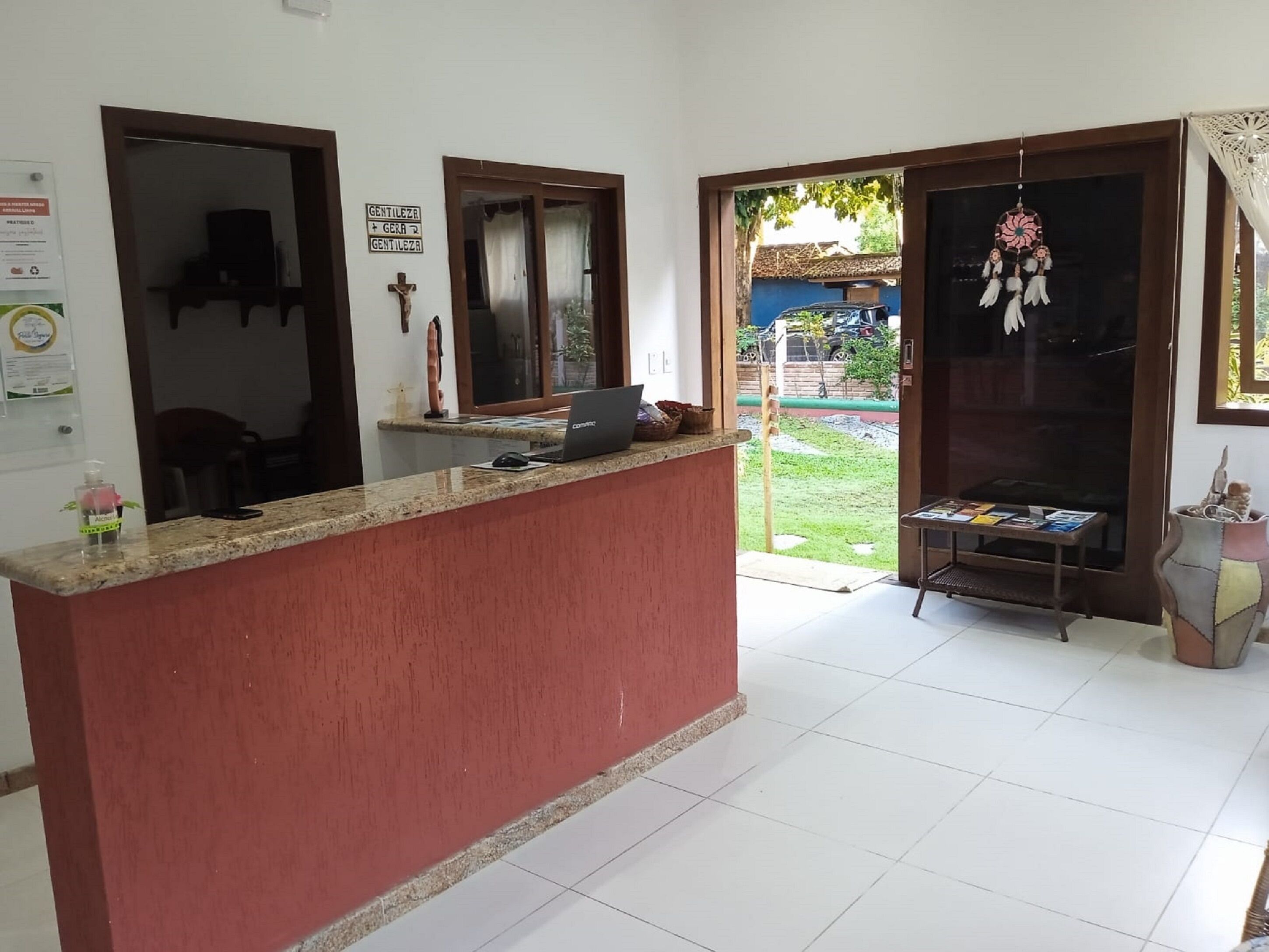 Zona de recepción con un mostrador rojo, una computadora portátil y una pequeña mesa con revistas. Hay dos ventanas con cortinas, una cruz en la pared y un atrapasueños colgando a la derecha.