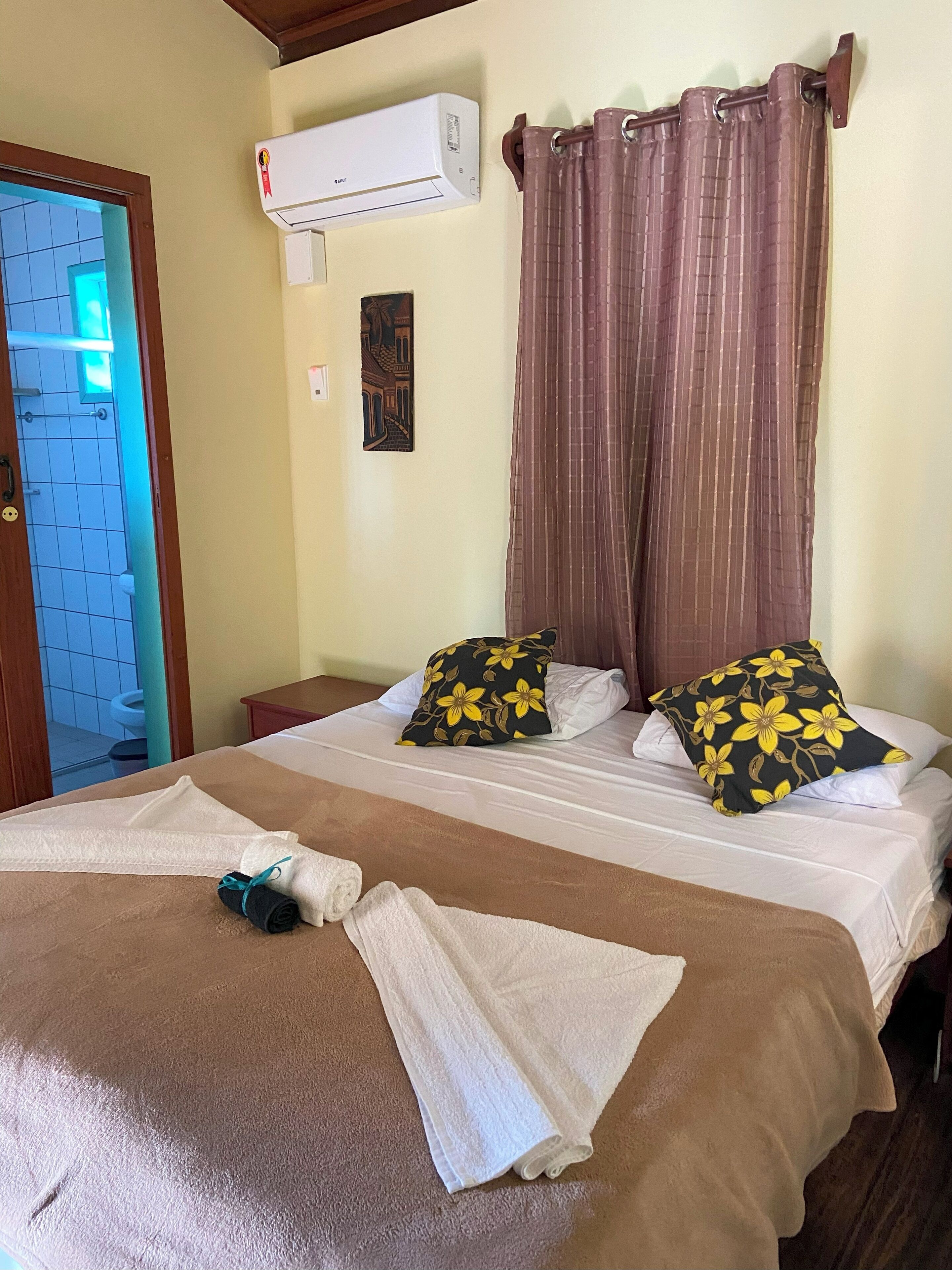 A hotel room with a bed, two pillows with yellow floral patterns, a towel, an air conditioner, a curtain, and a picture on the wall.