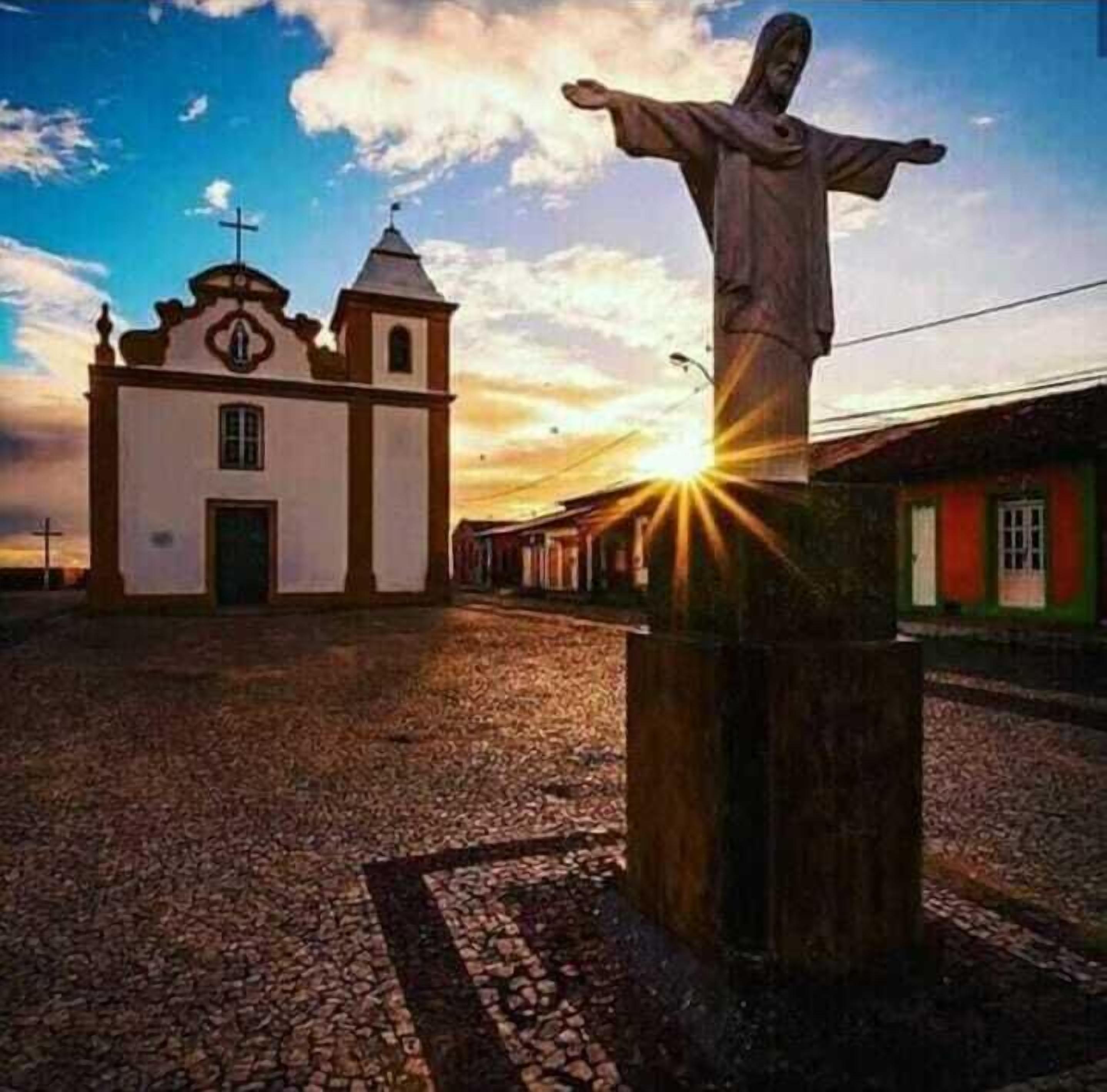 A church with a cross on top, a statue of a figure with outstretched arms, and a cobblestone ground.