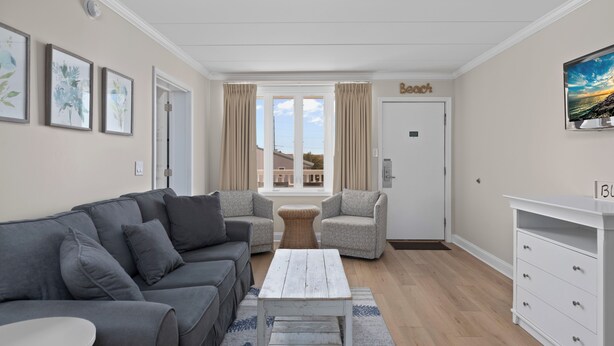 Ein Wohnzimmer mit grauem Sofa, einem weißen Couchtisch, Korbstühlen, einer weißen Kommode und einem großen Fenster mit Vorhängen.