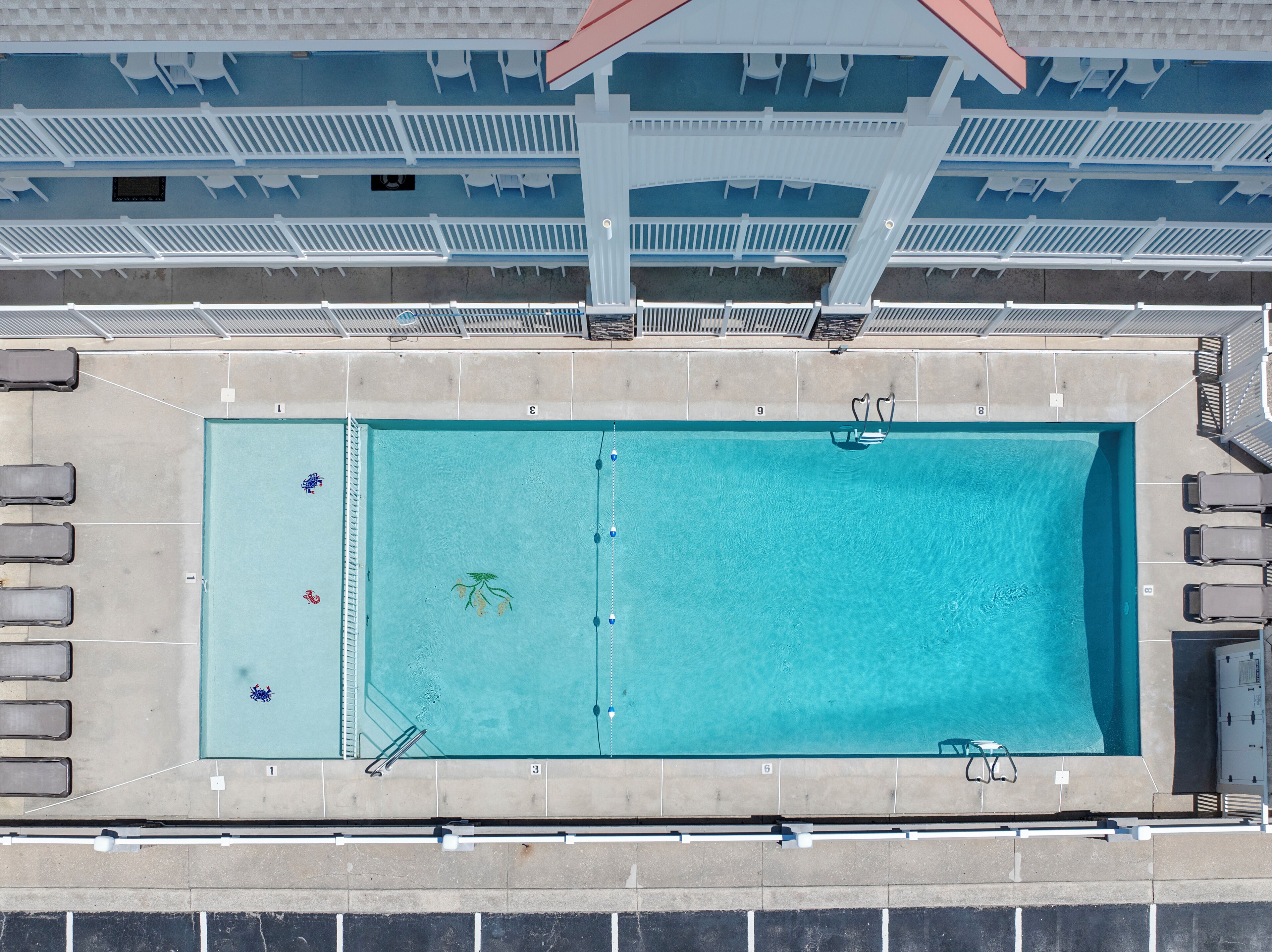 An aerial view of a rectangular swimming pool with a diving board, surrounded by lounge chairs and a covered seating area with white chairs.