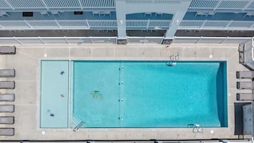 An aerial view of a rectangular swimming pool with a diving board, surrounded by lounge chairs and a covered seating area with white chairs.