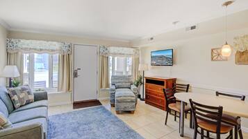 A cozy living room with a sofa, a dining table with chairs, a TV mounted on the wall, and a window with curtains.