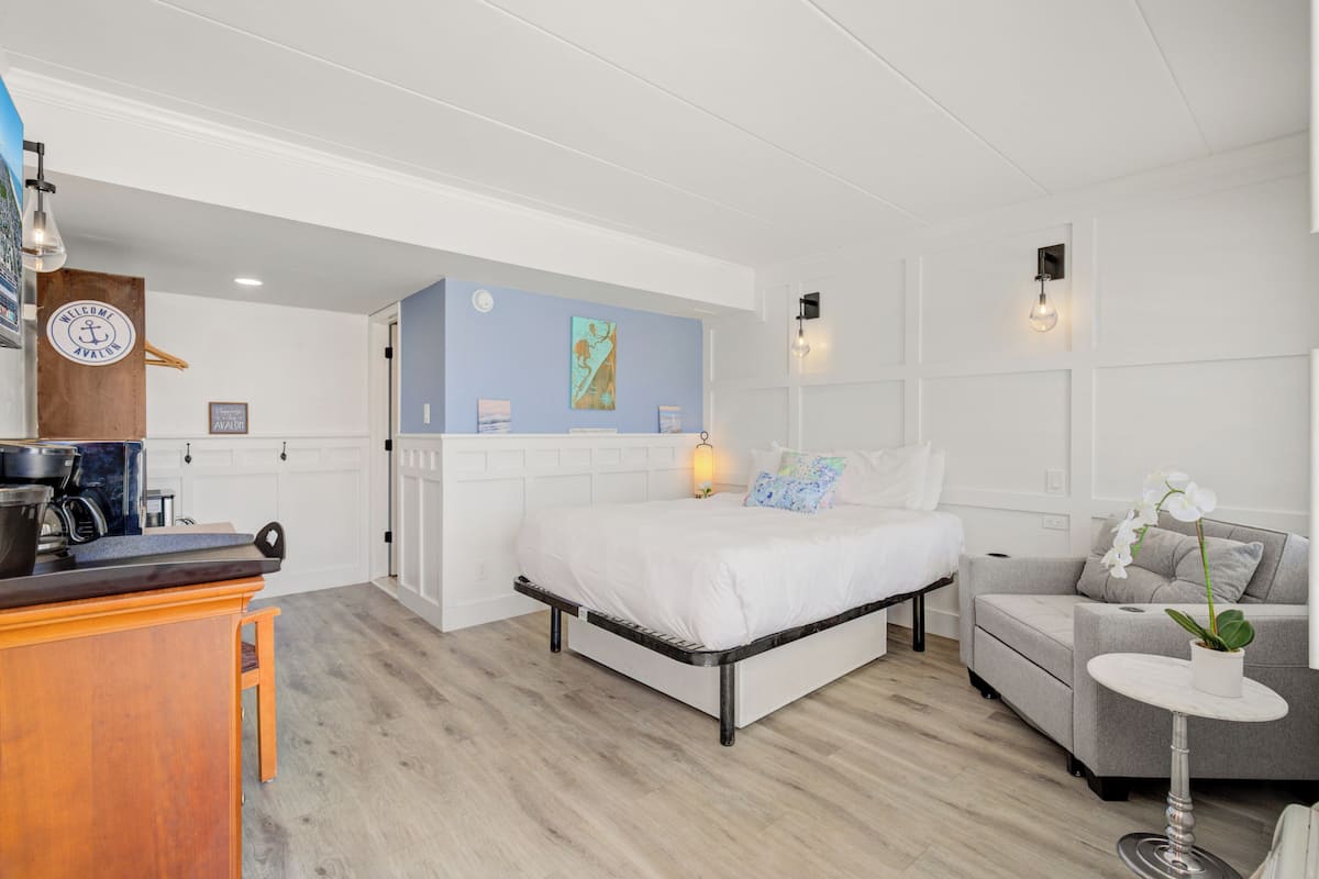 A bedroom with a bed, a sofa, a small table with a plant, a kitchen area, and a wooden cabinet.