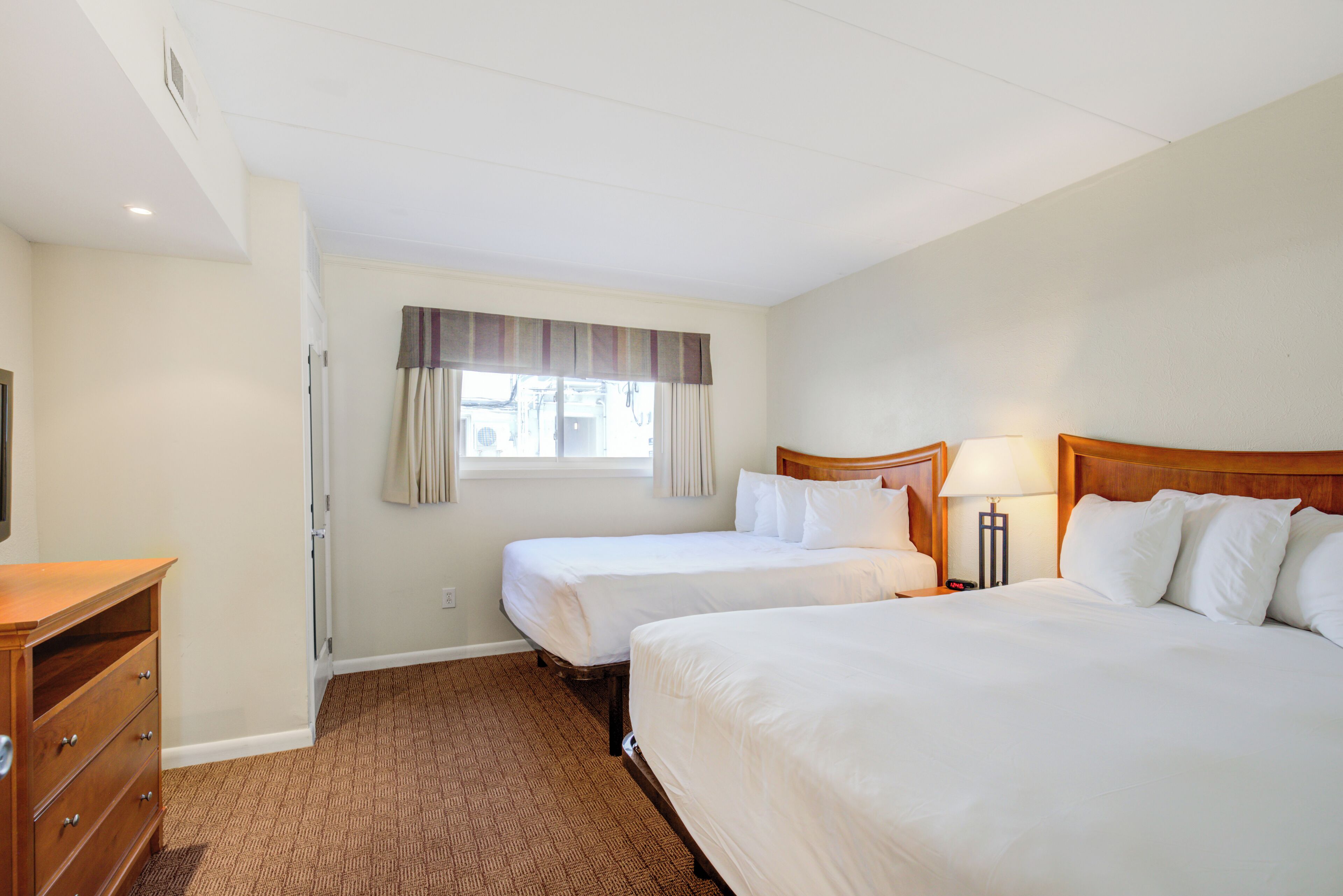 Une chambre d’hôtel avec deux lits, une commode en bois, une fenêtre avec des rideaux et une lampe sur la table de chevet.